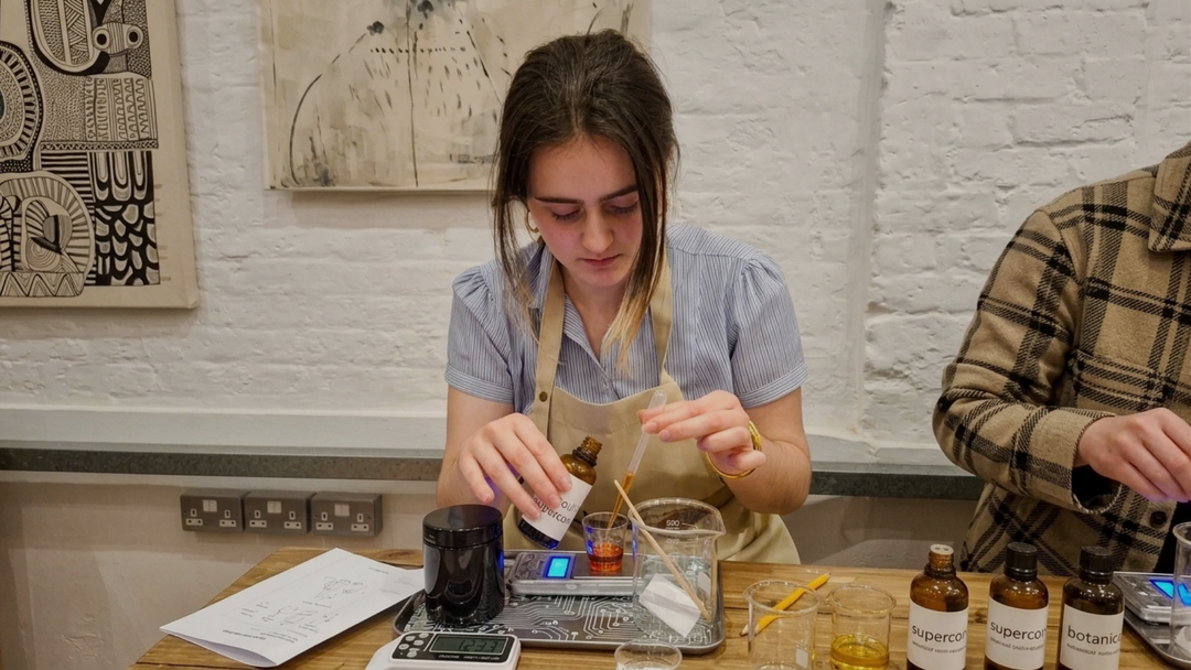 A woman wearing a light blue striped shirt and beige apron mixes ingredients in a glass beaker on a scale at a wooden table, with other people partially visible at the sides in a workshop setting.