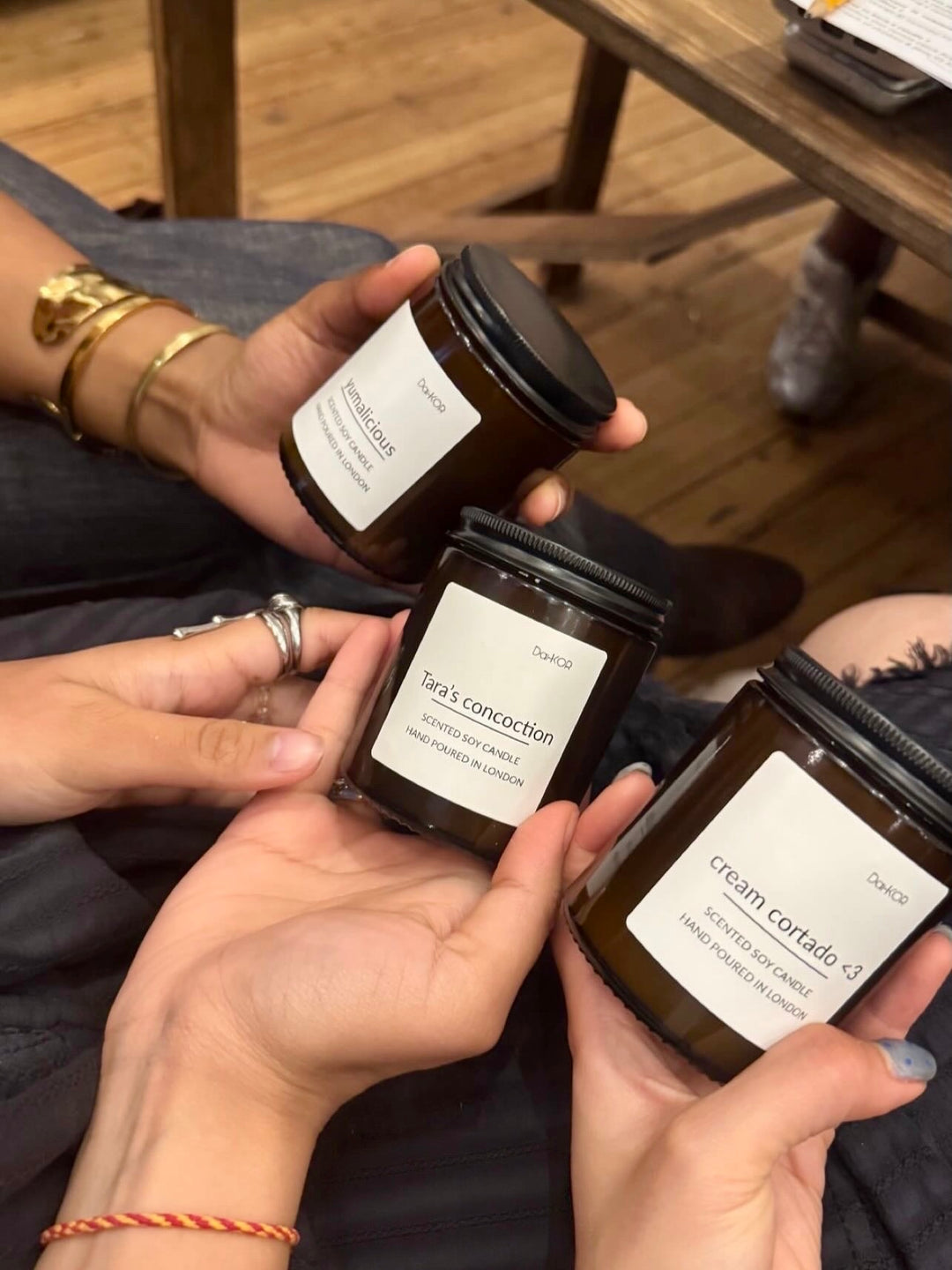 Three amber glass jar candles with white labels are held up by multiple hands in a casual indoor setting with a wooden floor and table visible in the background.